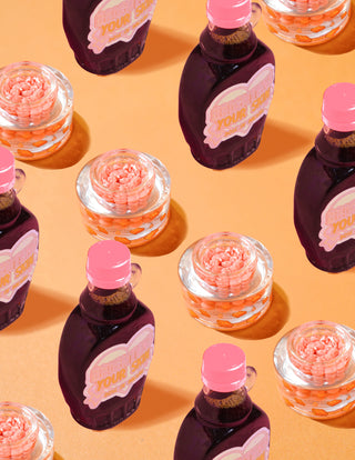 Clear containers of P.Louise Dose of Dreams Vitamin C Capsule Cream with orange capsules within them surrounded by purple and pink Syrup bottles on an orange background