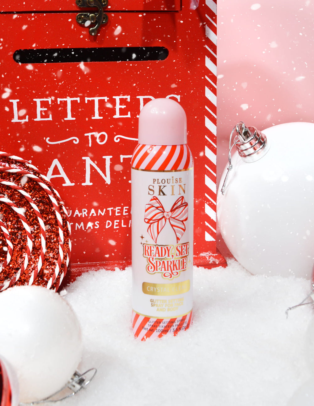 Butelka P. Louise Ready Set Sparkle Christmas Glitter Setting Spray otoczona świątecznymi dekoracjami, takimi jak śnieg, bombki i świąteczna skrzynka na listy