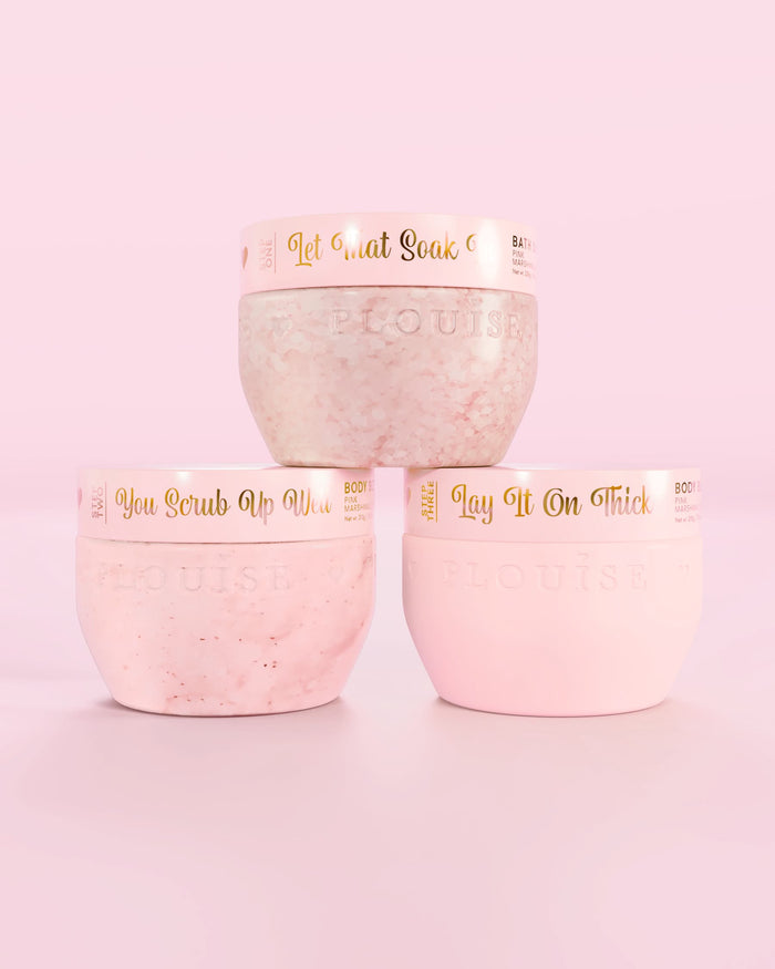 Trzy różowe pojemniki P.Louise Hop Into Self-Care Pink Marshmallow zawierające sole do kąpieli, peeling do ciała i masło do ciała ze złotym tekstem na różowym tle