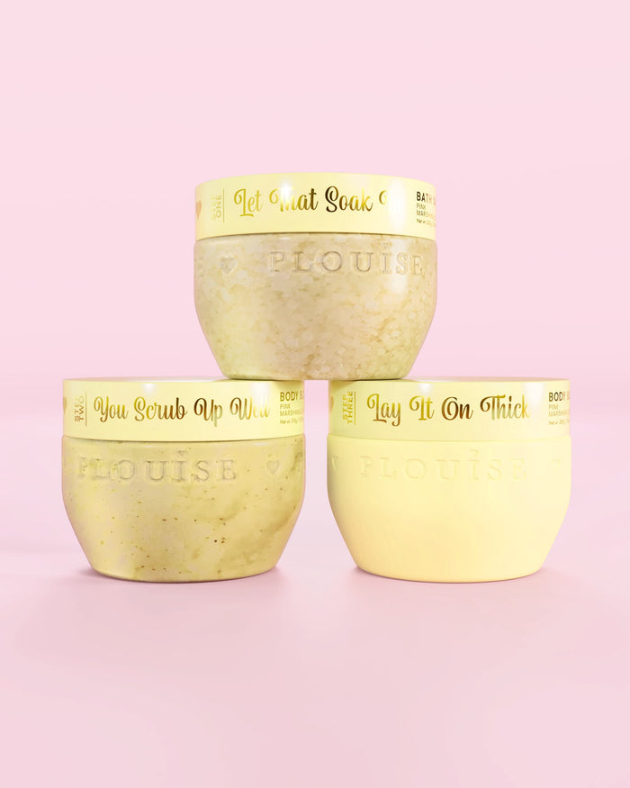 Trzy żółte pojemniki P.Louise Hop Into Self-Care Starwberry Shortcake zawierające sole do kąpieli, peeling do ciała i masło do ciała ze złotym tekstem na różowym tle