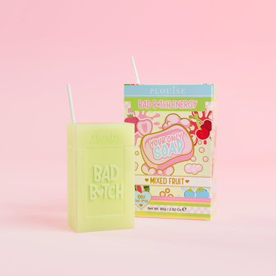Zielone mydło w kostce z wygrawerowanym napisem „Bad Bitch”, któremu towarzyszy kolorowe pudełko po soku z etykietą „Mixed Fruit”, prezentowane na różowym tle.