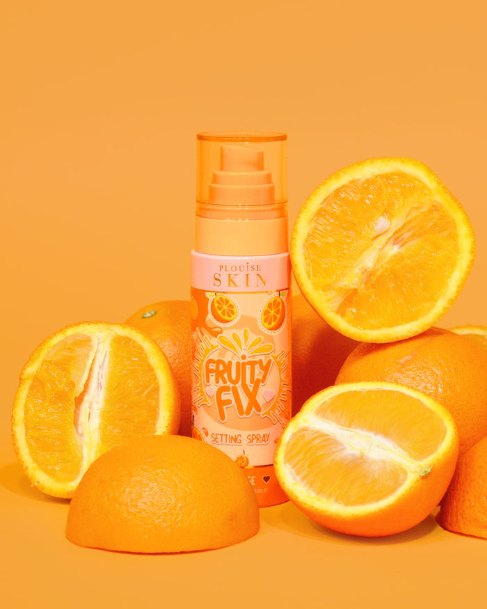 Pomarańczowa butelka sprayu utrwalającego witaminę C P. Louise Fruity Fix z pomarańczowym tłem otoczonym świeżymi plasterkami pomarańczy.